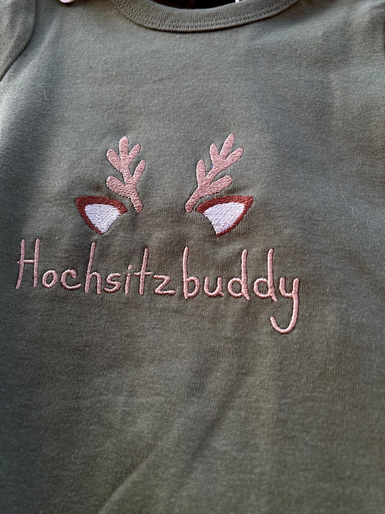 Puede incluir: Una camiseta verde de manga larga con cuernos de reno bordados en rosa y el texto "Hochsitzbuddy".