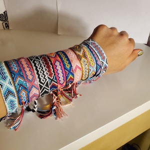 May include: A stack of colourful friendship bracelets with tassels. The bracelets are made of woven string and feature geometric patterns in shades of pink, blue, yellow, white and black.