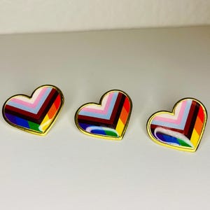 May include: Three heart-shaped pins with a gold border. Each pin features a colourful, geometric design with rainbow stripes. The pins are arranged in a row, showcasing their vibrant and eye-catching appearance.
