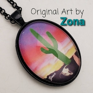 May include: Black oval pendant necklace featuring a desert landscape with a green cactus against a sunset sky. The pendant has a black frame and chain. The text "Original Art by Zona" is visible.