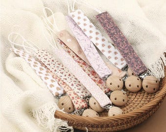 Könnte beinhalten: Eine Sammlung von Schnullerhaltern für Babys mit Holzperlen und Stoffbändern mit Mustern. Die Bänder weisen verschiedene Designs auf, darunter Regenbogen und Blumenmuster. Die Schnullerhalter sind in einem Weidenkorb angeordnet.