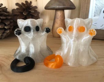 May include: Two translucent cat ghost figurines with black and orange accents. One has black eyes and paws, the other has orange eyes and paws. A black curved object and an orange ring are in front of them. Halloween decor.