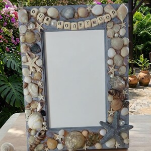 May include: A rectangular photo frame with a grey border, decorated with seashells, starfish, and pearls. The frame has the words "OUR HONEYMOON" spelled out in individual wooden letters. A large blue starfish adorns the bottom right corner.