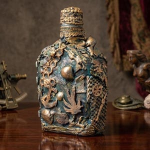 May include: A decorative glass bottle featuring a textured, dark blue and bronze finish. The surface is adorned with various nautical-themed embellishments, including a metal anchor, seashells, a ship wheel, and netting, all finished in a metallic bronze.