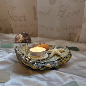 May include: A decorative oyster shell candle holder featuring a lit white tea light candle, a small beige starfish, and tiny decorative accents. The shell has a textured, dark, and metallic finish, resting on a smooth, light-coloured fabric surface.