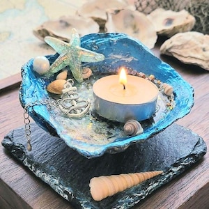 May include: A blue painted oyster shell candle holder containing a lit tea light candle, a small silver anchor charm on a chain, a glittery starfish, and several tiny seashells, all resting on a square black slate coaster.