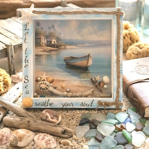 May include: A framed coastal art piece depicting a tranquil beach scene with a boat, and the text "Let the Sea soothe your soul." The light blue frame is decorated with rope and seashells. Seashells and sea glass are scattered around the frame.