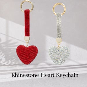 Pode incluir: Dois chaveiros de coração de strass, um vermelho e outro transparente. Ambos os chaveiros têm um anel dourado e um pingente de coração dourado.
