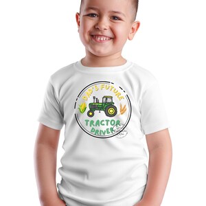 May include: White short-sleeved t-shirt featuring a circular graphic with a green tractor, corn, and wheat. The text reads "DAD'S FUTURE TRACTOR DRIVER". The design is in a cartoon style, suitable for children.