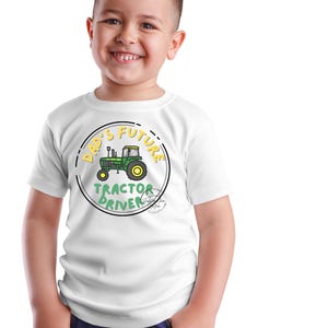 May include: A white t-shirt featuring a green and yellow tractor graphic with the text "DAD'S FUTURE TRACTOR DRIVER" in a circular design. The shirt is worn by a smiling child wearing blue jeans.
