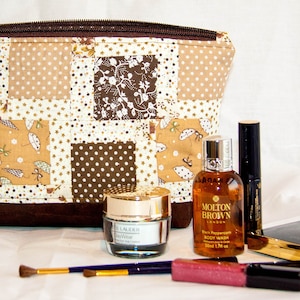 Può includere: Una trousse da toilette imbottita di colore marrone con cerniera e diversi motivi marroni e bianchi. La trousse è piena di articoli da toeletta, tra cui una bottiglia di bagnoschiuma, un barattolo di crema per il viso, un tubo di lucidalabbra e un mascara. La trousse è appoggiata su una superficie bianca.