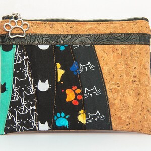 May include: A rectangular zippered pouch with a cat theme. The pouch features a patchwork design with a cork panel, black fabric with white cat faces and colorful paw prints, and a teal panel. A silver paw print charm hangs from the zipper.