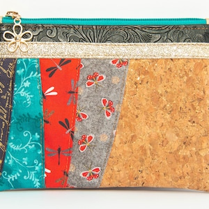 Peut inclure: Une petite pochette zippée avec un extérieur en liège et un design patchwork. Le patchwork comprend du tissu turquoise, rouge et gris avec des motifs de libellules et de fleurs. La pochette a un tirette de fermeture éclair dorée avec un charme de fleur décoratif et une étiquette noire qui indique "Handmade by Magic Flying Boots".