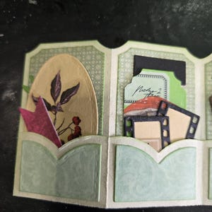 May include: A handmade paper craft with multiple pockets. The pockets are decorated with light green and white patterned paper. One pocket contains a pink paper arrow and a tan oval with a botanical illustration. Other pockets hold film strips and tags.