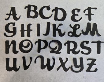 May include: Black, die-cut uppercase alphabet letters arranged on a white surface. The letters are in a cursive font, with a variety of swirls and curves. The letters are evenly spaced and clearly visible.