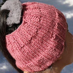 May include: A pink knitted hat with a hole in the back for a ponytail. The hat is made with a textured stitch and has a gray fuzzy scrunchie holding the ponytail.