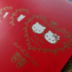 May include: Red wedding envelopes with gold accents featuring Hello Kitty and Daniel. The envelopes have a gold heart design with the text "Hello Kitty & Dear Daniel" and a Chinese character for double happiness.