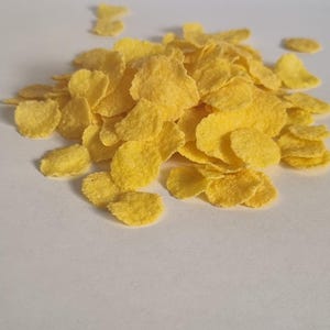 Könnte beinhalten: Ein Haufen gelber Cornflakes auf weißem Hintergrund.