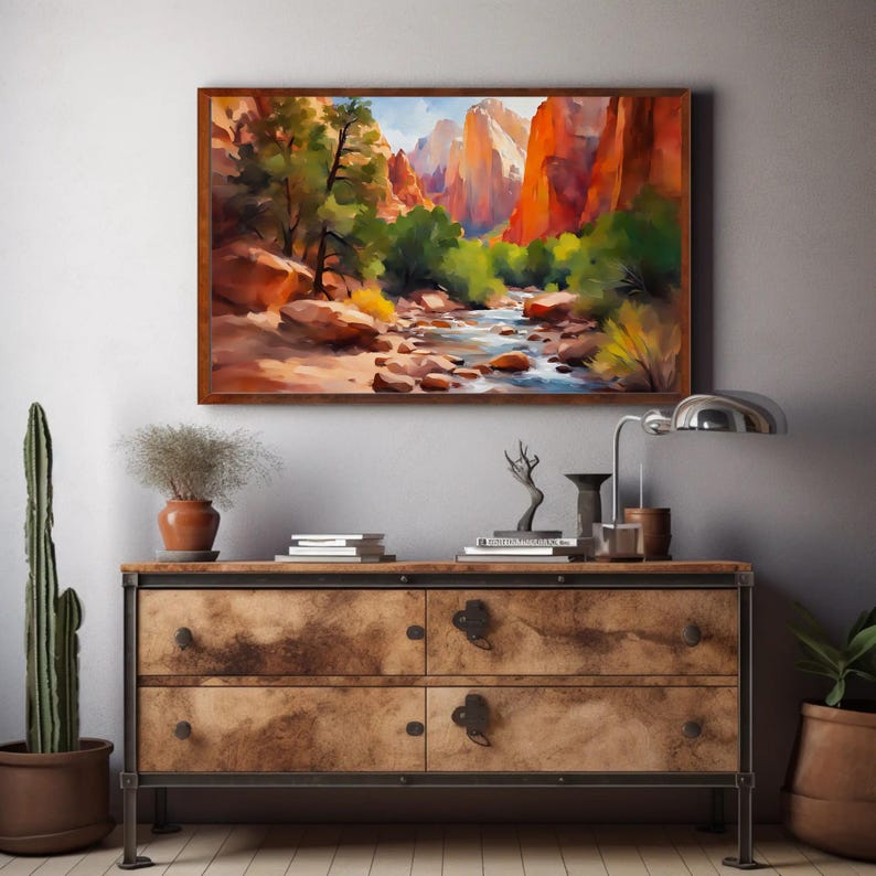 May include: A framed landscape painting depicts a canyon scene with a river, trees, and red rock formations. The artwork is displayed above a wooden dresser with metal accents, alongside decorative items and a potted cactus.