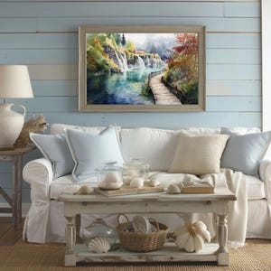 May include: A framed watercolor painting of a waterfall scene hangs above a white sofa with light blue pillows. A wooden coffee table displays decorative shells, a basket, and a white pumpkin. A lamp and side table are on the left.