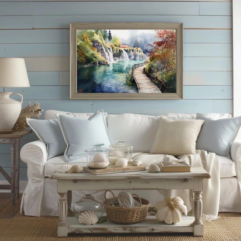 May include: A framed watercolor painting of a waterfall scene hangs above a white sofa with light blue pillows. A wooden coffee table displays decorative shells, a basket, and a white pumpkin. A lamp and side table are on the left.