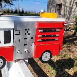 May include: A handmade mailbox designed as a fire truck. The mailbox is red, white, and silver with black and silver wheels. It has a red flag and a yellow bar on top. The mailbox is mounted on a white post.