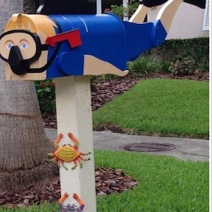 May include: A novelty mailbox shaped like a scuba diver wearing a blue wetsuit, black mask, and fins, mounted on a white post. The post features two decorative crab figures, one yellow and one purple, attached to the front surface below the mailbox.