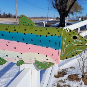 May include: A whimsical, hand-painted mailbox shaped like a fish. The fish-shaped mailbox is painted with green, blue, pink, and white stripes, and black spots. It is mounted on a white fence in a snowy outdoor setting.