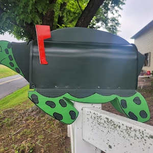 May include: A dark green mailbox shaped like a turtle, with a red flag. The turtle design includes green and black details on the head and feet. The mailbox is mounted on a white fence.