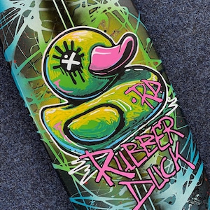 Könnte beinhalten: Ein schwarzes Skateboard-Deck mit einer Illustration einer grünen, gelben und pinken Gummi-Ente und den Worten "Rubber Duck" in pinker Farbe.