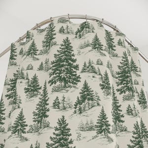 May include: A shower curtain featuring a repeating forest pattern with dark green pine trees printed on a light cream background. The fabric hangs from a curved metal rod with silver-toned rings, displaying a detailed winter landscape design.