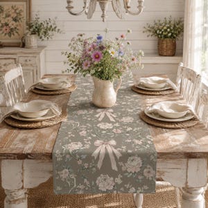 Puede incluir: Una mesa de comedor preparada para una comida, con un camino de mesa con estampado floral y un diseño de lazo. La mesa está puesta con platos blancos, manteles individuales tejidos y un jarrón con flores. La mesa es de madera y tiene sillas blancas.