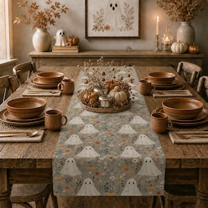 May include: A rustic wooden dining table set for a fall-themed dinner. The table is adorned with a gray table runner featuring a pattern of smiling ghost figures and floral accents. Brown ceramic dishes and mugs are set for each place setting.
