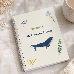 Pode incluir: Um planejador de gravidez com espiral com o nome "Victoria" e o título "My Pregnancy Planner" em verde e azul. A capa apresenta uma ilustração de baleia azul e detalhes botânicos. Uma caneca e um cobertor dobrado estão no fundo.