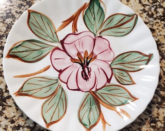 May include: White ceramic plate with a scalloped edge, featuring a hand-painted floral design. The central flower is pink with brown and yellow details, surrounded by green leaves and brown stems. A decorative piece for serving or display.
