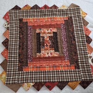 May include: A square log cabin quilt block featuring a central arrangement of fabric strips in shades of orange, brown, and cream. The design includes various patterns like floral, plaid, and leaf prints, finished with a wide brown plaid border.