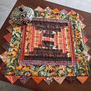 May include: A square fabric quilt mat featuring a central log cabin pattern in shades of brown and orange, surrounded by a border of pumpkins and gourds. A round, lidded ceramic jar with a blue, green, and yellow lattice pattern sits on top.