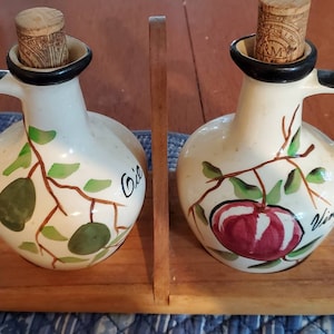 May include: A set of two ceramic cruet bottles for oil and vinegar, with cork stoppers. The bottles are white with black handles and rims, and feature hand-painted olive and apple designs. The bottles sit in a wooden holder.