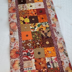 May include: A rectangular patchwork table runner with a fall theme. It features squares of various fabrics in orange, brown, and yellow, adorned with wooden buttons. The edges are bordered with a pumpkin and leaf print.