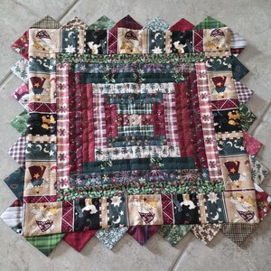 May include: A square fabric quilt block featuring a log cabin pattern with concentric borders of red, green, and tan fabric strips. The outer border displays a repeating print of angels holding stars, interspersed with moon and star motifs.
