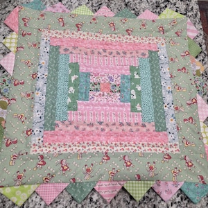May include: A handmade quilt with a log cabin design. The quilt features a central square of pink and floral fabrics, surrounded by concentric rectangles of various patterns and colors, including pink, green, and blue. The border is adorned with alternating pink and green triangles.