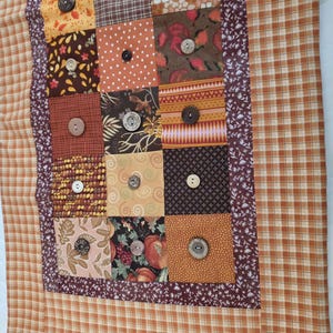 May include: A rectangular fabric table runner featuring a patchwork design of various autumn-themed patterns in shades of orange, brown, and cream. The center panel consists of square fabric blocks, each embellished with a single round button.
