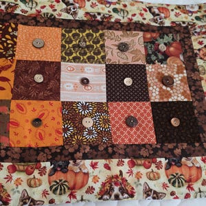 May include: A rectangular fabric quilt featuring a patchwork center of brown, orange, and cream squares, each adorned with a single round button. The border displays a repeating pattern of puppies, pumpkins, and autumn leaves in warm seasonal tones.