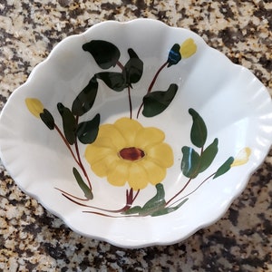 May include: A white ceramic bowl with a scalloped edge features a yellow flower with brown center and green leaves. The bowl is decorated with a floral pattern.