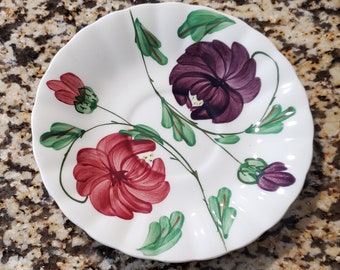 May include: White ceramic saucer with a scalloped edge, featuring a hand-painted floral design. The design includes two pink and purple flowers with green leaves and stems.