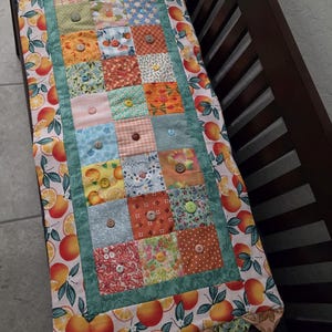May include: A rectangular fabric table runner featuring a central patchwork design of various patterned squares in shades of orange, blue, green, and pink, each accented with a small button. The border is a white fabric with orange citrus fruit.