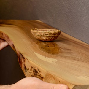 Könnte beinhalten: Ein Holzregal mit einer natürlichen Kante und einem geflochtenen Korb oben. Das Regal ist aus hellbraunem Holz mit dunkelbraunen Streifen. Der Korb ist aus Naturfasern und hellbraun.