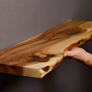 May include: A natural wood shelf with a live edge, showing the natural grain and texture of the wood. The shelf is being held up by two hands.