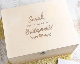 May include: A wooden box with the text "Sarah, Will You Be My Bridesmaid?" engraved on the lid. The box is closed and sitting on a gold glitter surface.