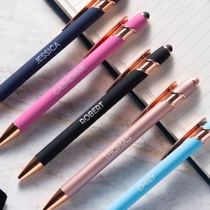 The image shows a collection of stylus pens laid out on a marble surface next to an open lined notebook. Each pen has a metallic finish, with what appears to be copper-colored tips and clips.